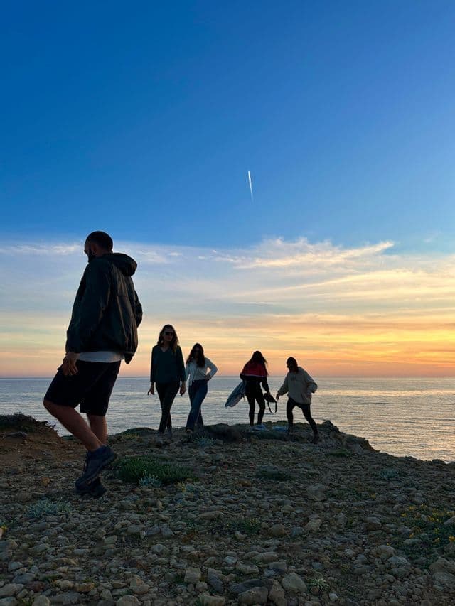 Un gruppo WeRoad in viaggio si trova su una scogliera rocciosa, ammirando il tramonto sull'oceano.