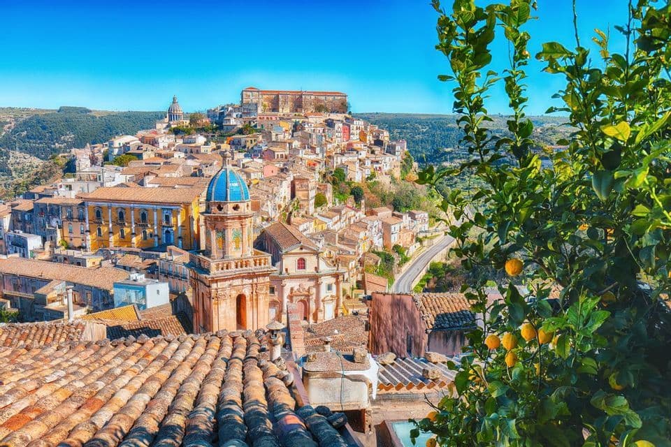 Una veduta soleggiata sui tetti di terracotta di un borgo collinare con una chiesa dalla cupola azzurra, incorniciata da un albero di limoni.