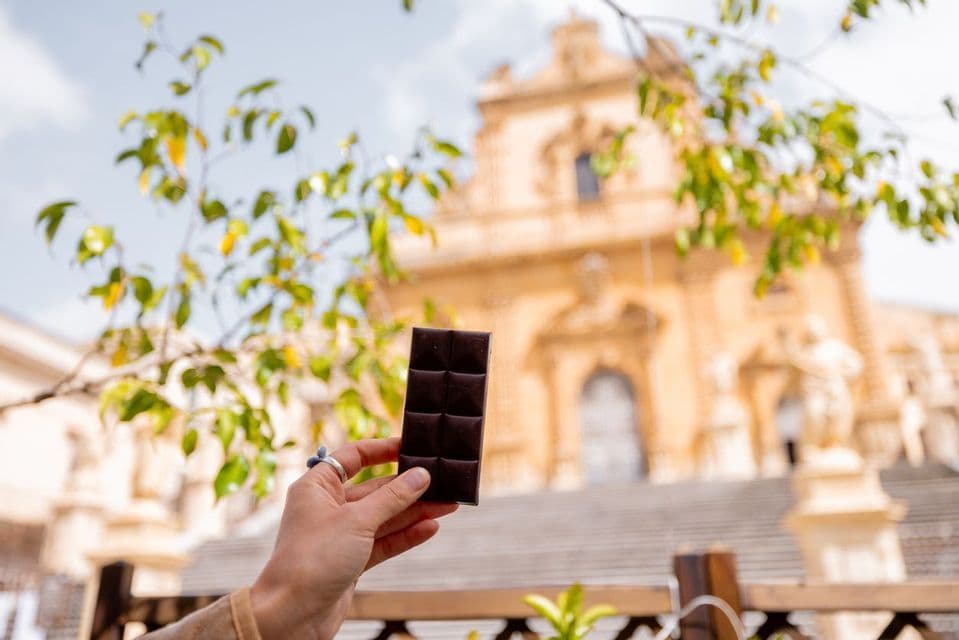 Una mano regge una tavoletta di cioccolato fondente con un grande edificio ornato sfocato sullo sfondo, sotto alcune foglie verdi.