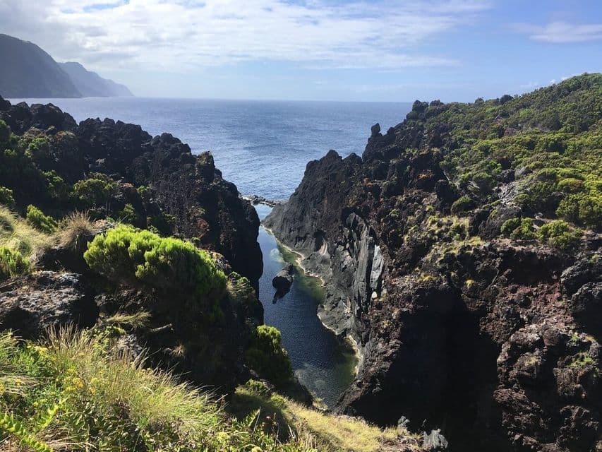 Eine schmale Bucht bildet einen natürlichen Pool zwischen dunklen, von grünen Sträuchern bewachsenen Vulkanfelsen, mit Blick auf den tiefblauen Ozean.