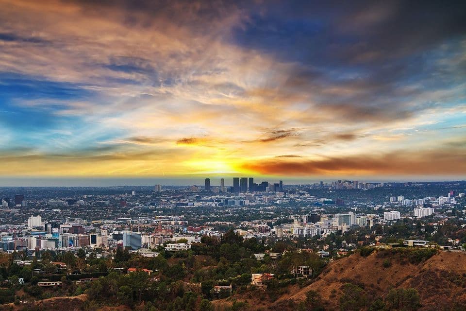A panoramic view of a sprawling city skyline with skyscrapers, seen from a hill during a colorful sunset.