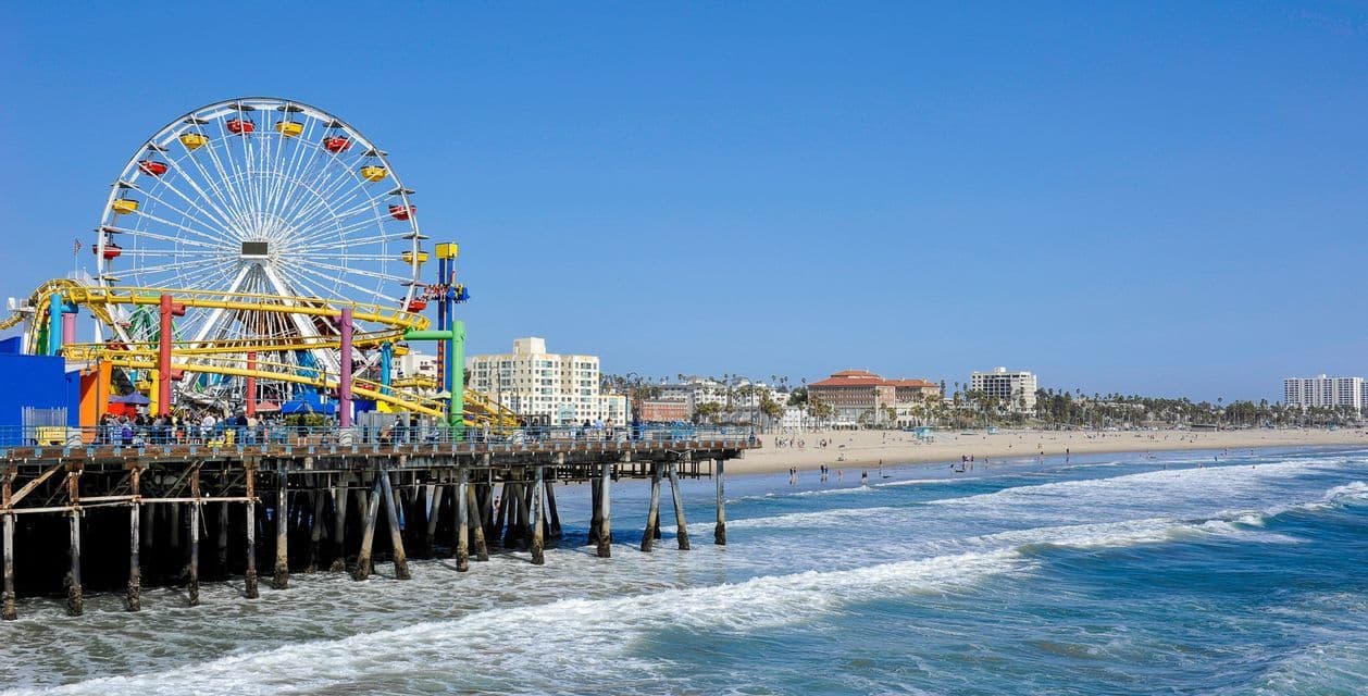 A pier with a large Ferris wheel and amusement park rides extends over the ocean next to a sandy beach under a clear blue sky.