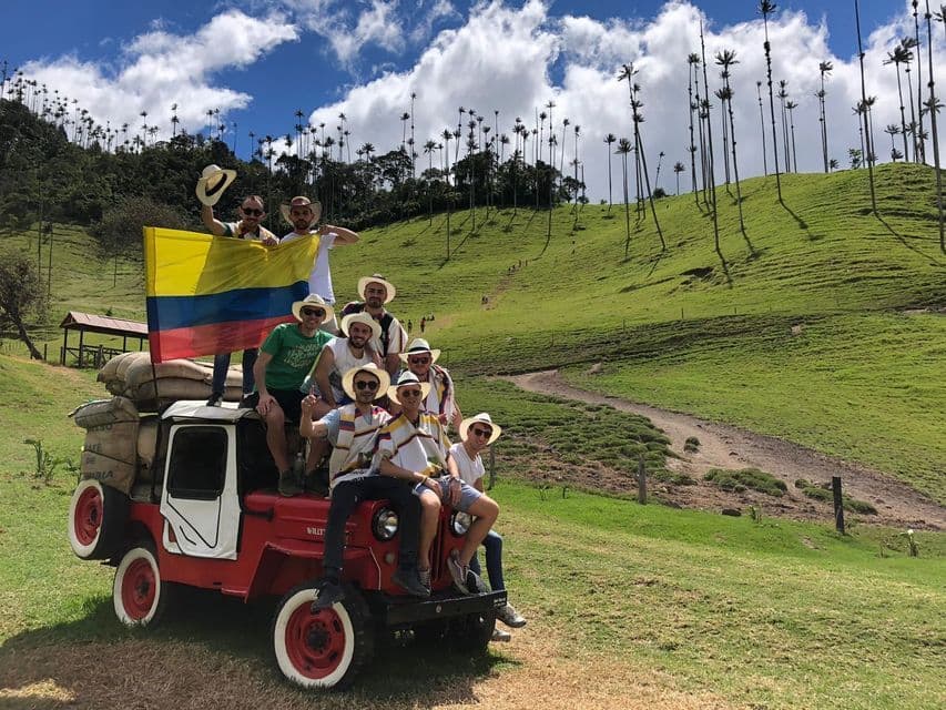 Un groupe WeRoad pose sur une jeep rouge, drapeau colombien en main, dans une vallée verdoyante parsemée de grands palmiers.