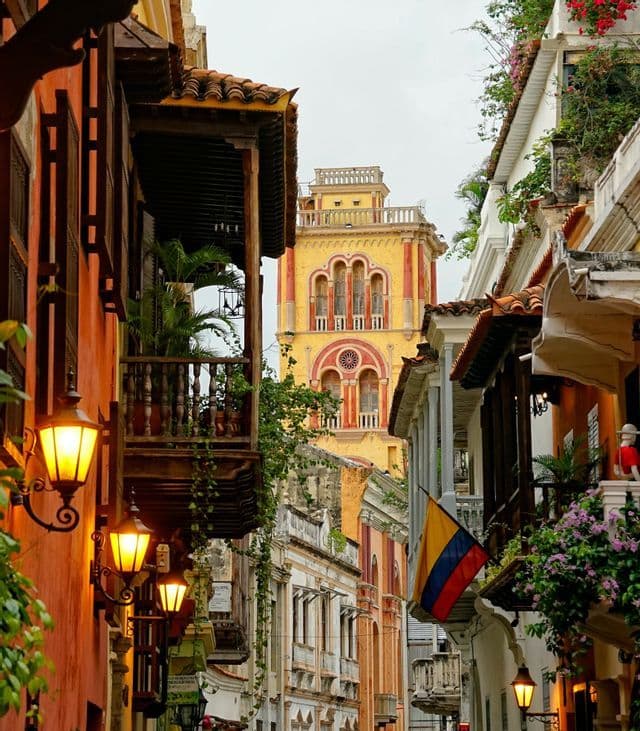 Una strada stretta con edifici colorati, lampioni accesi e una bandiera colombiana. Una torre dell'orologio gialla è visibile in lontananza.