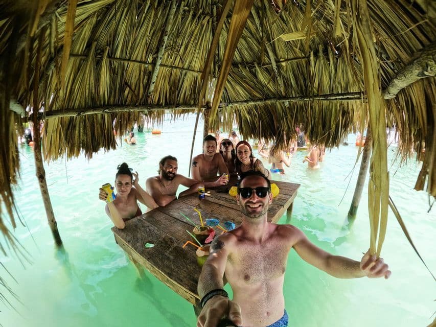 Un gruppo WeRoad scatta un selfie seduto a un tavolo di legno immerso nell'acqua turchese, sotto una capanna dal tetto di paglia.