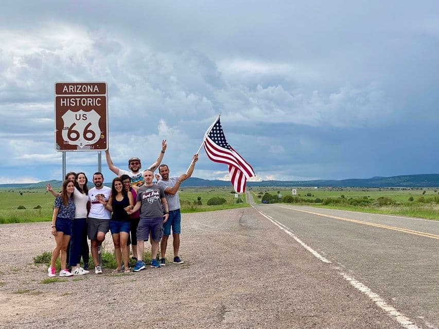 Eine WeRoad-Gruppe posiert an einem historischen US Route 66 Schild in Arizona, wobei eine Person eine amerikanische Flagge neben einer ländlichen Straße hält.