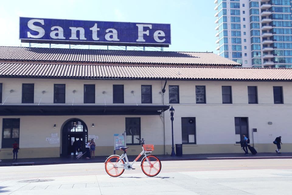 Una bicicletta arancione e bianca è parcheggiata su un marciapiede cittadino di fronte all'edificio storico del Santa Fe Depot in una giornata di sole.