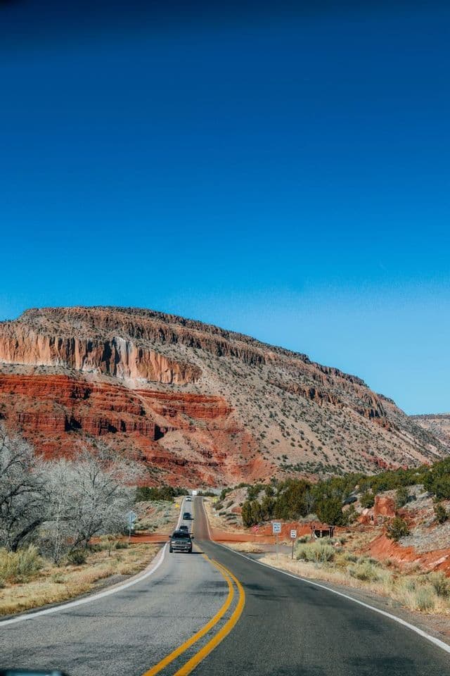 Des voitures roulent sur une route sinueuse, longeant une grande montagne de roche rouge stratifiée sous un ciel bleu éclatant.