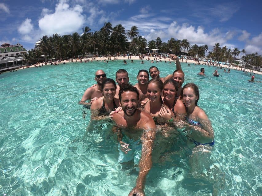Un gruppo WeRoad in viaggio si scatta un selfie sorridendo nell'acqua limpida e turchese dell'oceano, con una spiaggia affollata e palme sullo sfondo.