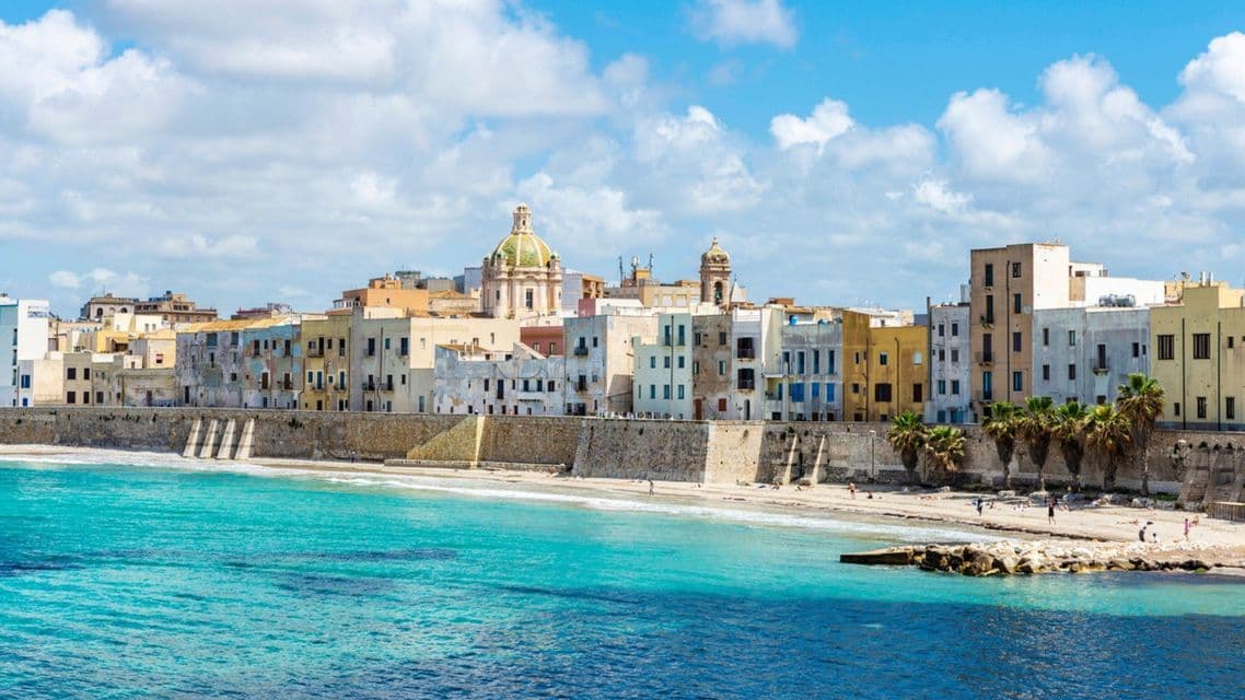 Un paesaggio urbano di edifici colorati lungo una diga di pietra, accanto a una spiaggia sabbiosa e acque turchesi.