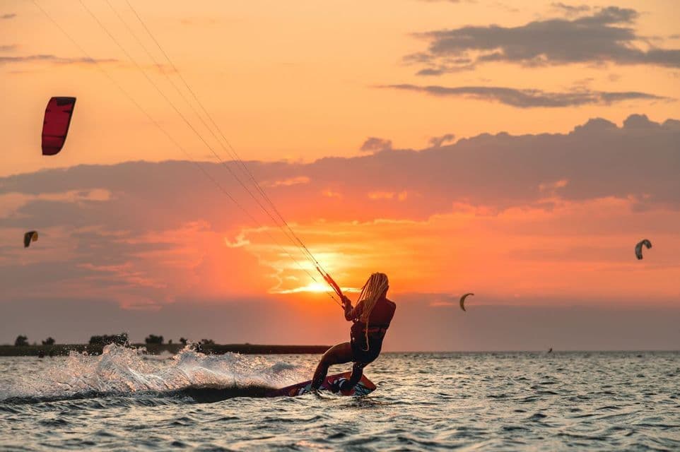 Una persona con i capelli lunghi fa kitesurf sull'acqua, stagliata contro un vibrante tramonto arancione.