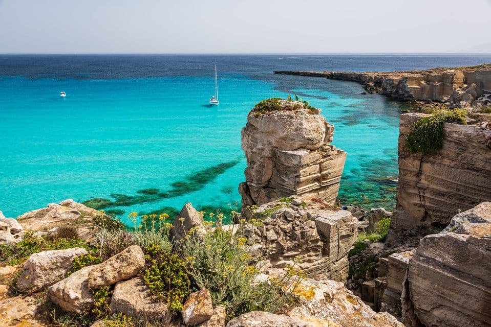 Una vista da una scogliera rocciosa che si affaccia su una cala con acqua turchese, dove una barca a vela è ancorata sotto un cielo sereno.