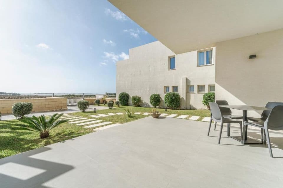 Un patio arredato con tavolo e sedie accanto al prato di un edificio moderno, con pietre calpestabili e cespugli, in una giornata di sole.