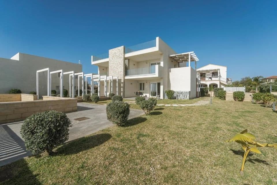 Un moderno edificio bianco a due piani con un prato all'inglese, cespugli potati e un vialetto pavimentato sotto un cielo azzurro e sereno.