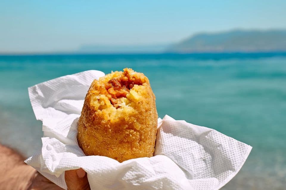 Una mano tiene un arancino fritto avvolto in un tovagliolo, con il mare azzurro brillante e una costa lontana sullo sfondo.