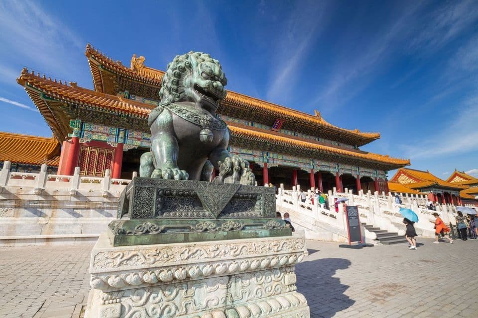 Une statue de lion gardien en bronze sur un piédestal, devant un palais chinois traditionnel, sous un ciel bleu clair.