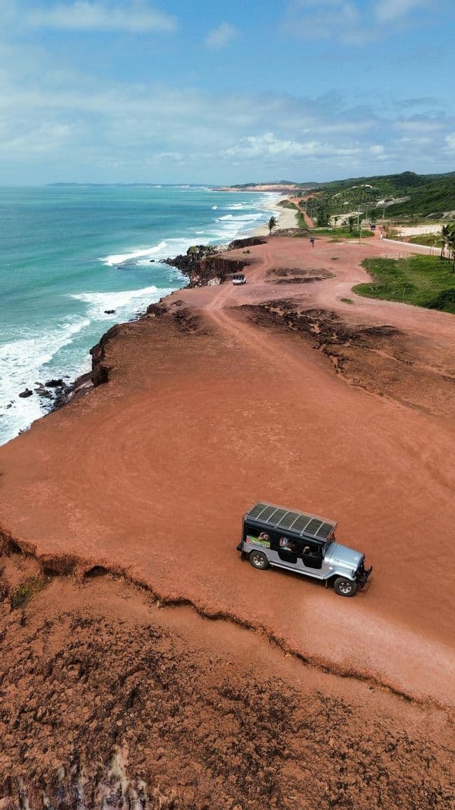 Vista aerea di un viaggio di gruppo WeRoad che guida un fuoristrada lungo un sentiero sulla scogliera rossa accanto all'oceano turchese.