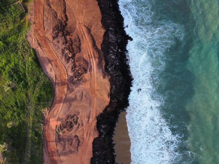 Vista aérea de un grupo WeRoad caminando por un sendero de tierra rojiza en la costa, donde la tierra se une al océano.