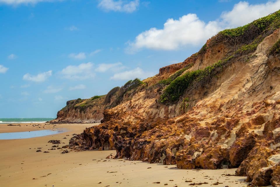 Rötlich-orange Klippen mit grüner Vegetation säumen einen Sandstrand unter einem teilweise bewölkten blauen Himmel.