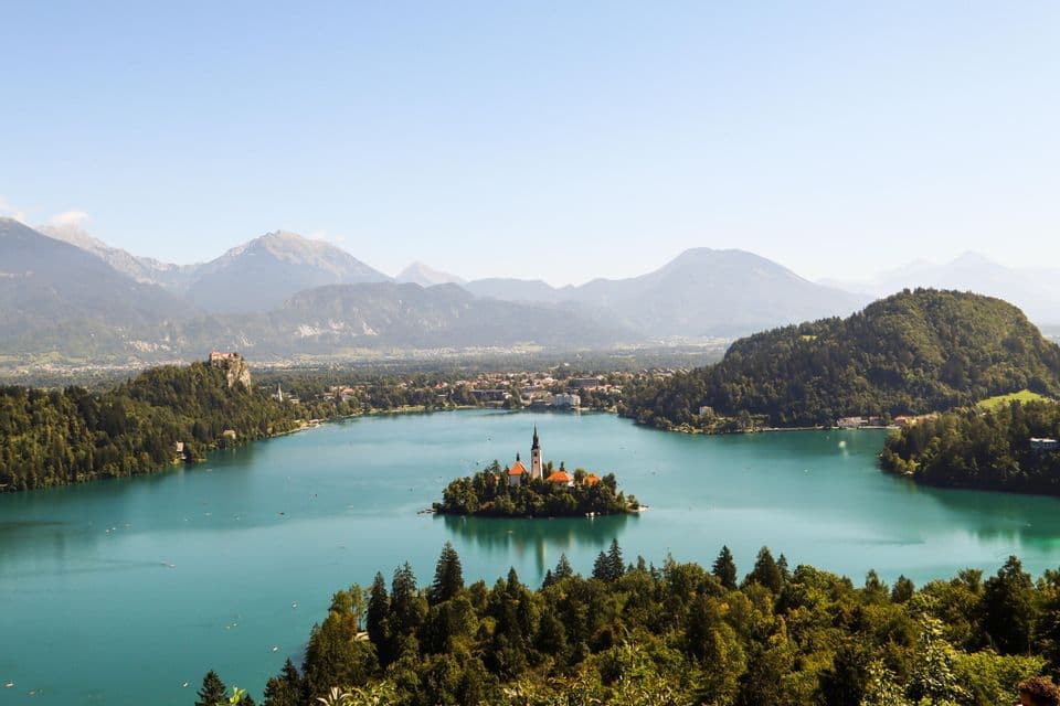 Una vista aerea di una chiesa su un'isola in un lago turchese, circondata da verdi foreste e montagne lontane.