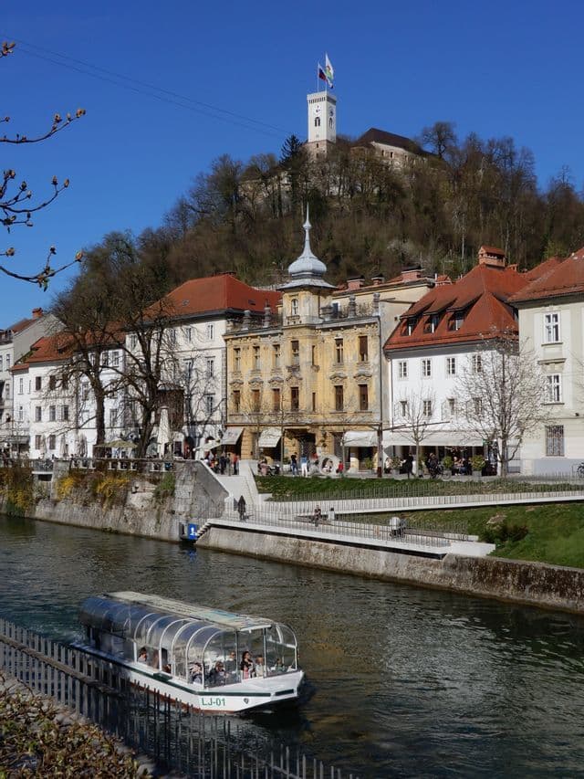 Un battello turistico con tetto in vetro naviga lungo un fiume, affiancando un lungofiume con edifici storici e un castello in cima a una collina sullo sfondo.