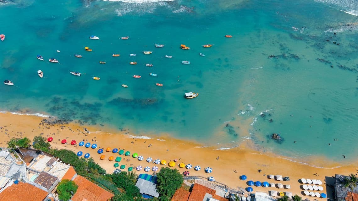 Una veduta aerea di una spiaggia sabbiosa con ombrelloni colorati e barche che galleggiano sul mare turchese.