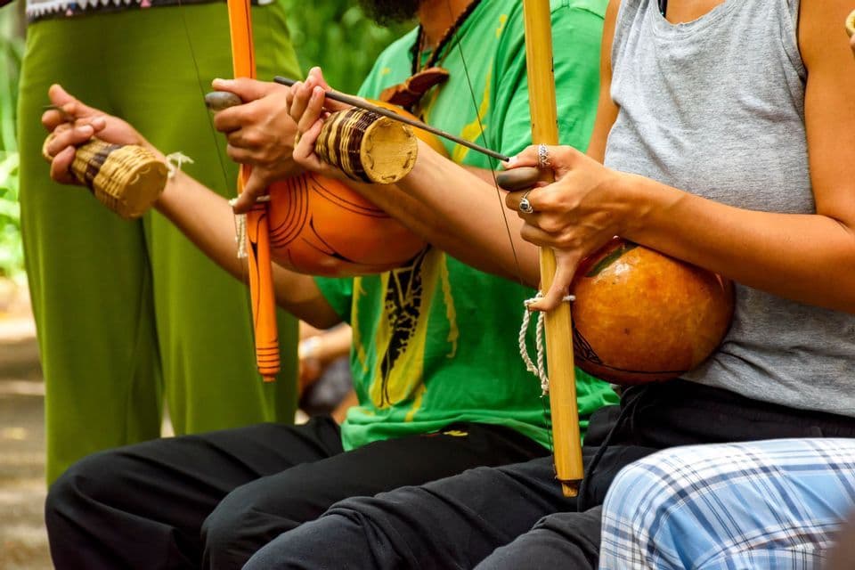 Un primo piano di tre persone sedute in fila, che suonano strumenti musicali tradizionali berimbau e caxixi.