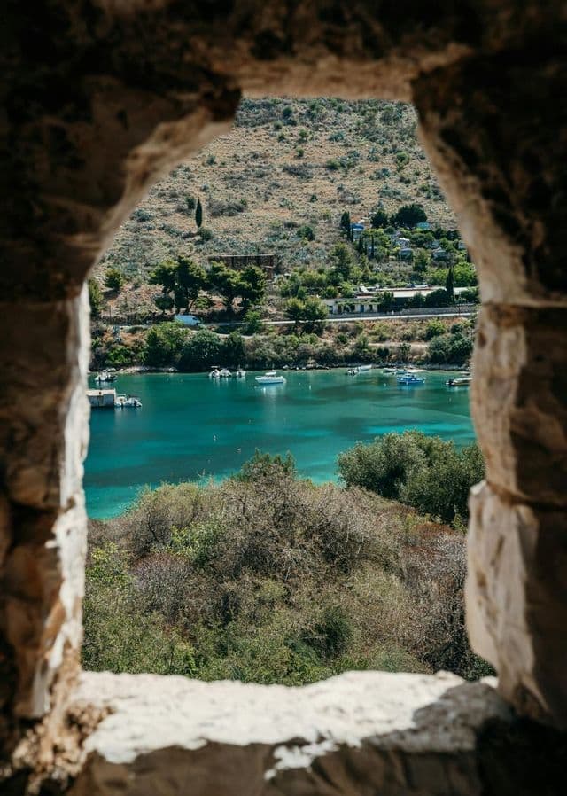 Una vista attraverso una finestra di pietra di una baia costiera con acqua turchese, piccole imbarcazioni e una collina con alberi verdi ed edifici.