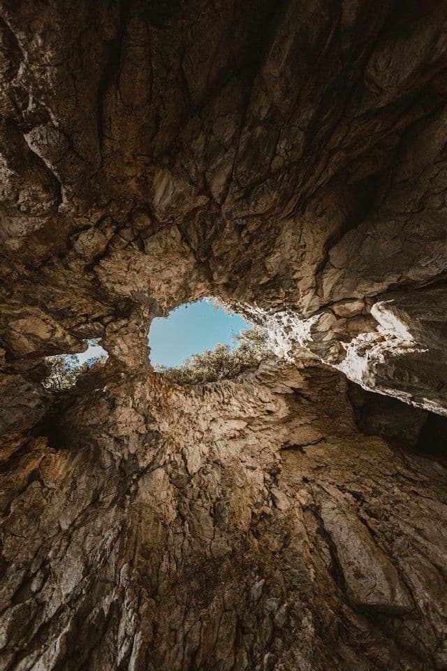 Vista dal basso dall'interno di una grotta rocciosa, con lo sguardo rivolto al cielo blu e alle cime degli alberi attraverso un'apertura naturale.