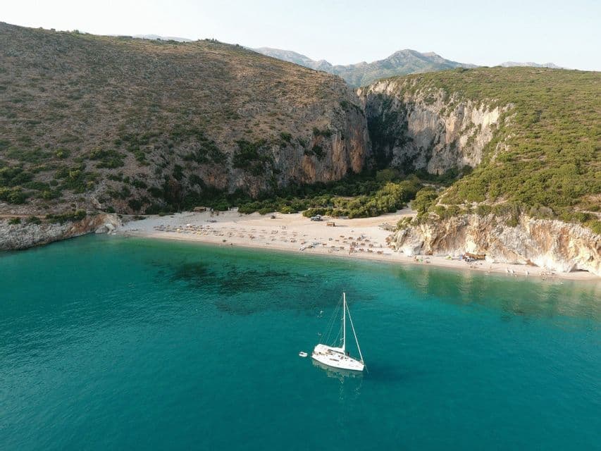 Veduta aerea di una barca a vela bianca ancorata in una baia turchese, con una spiaggia sabbiosa isolata incastonata tra ripide scogliere.