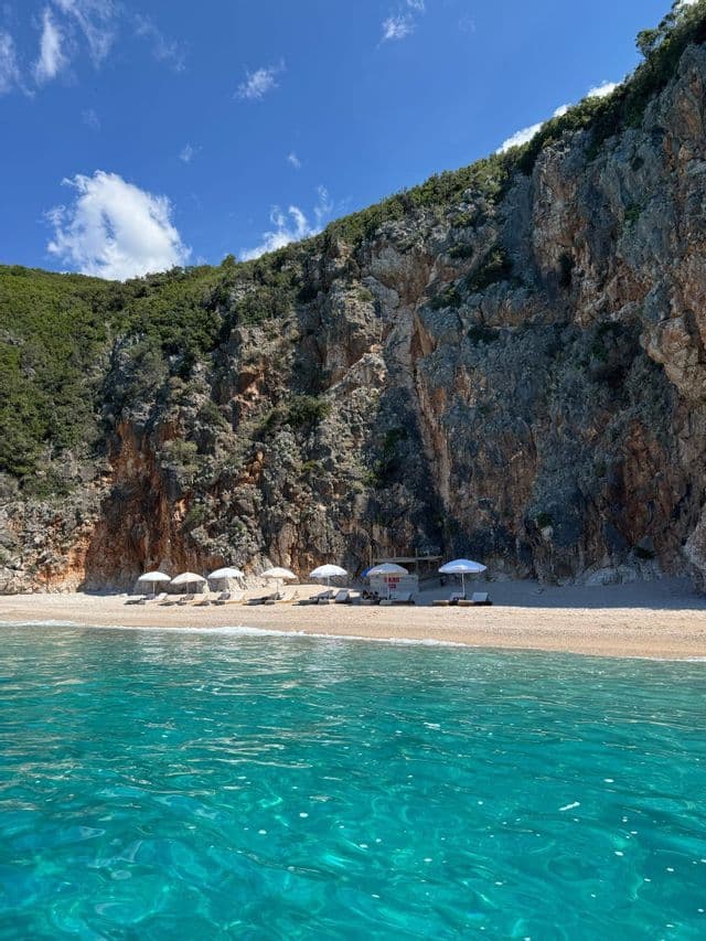Vista dall'acqua cristallina e turchese di una spiaggia sabbiosa appartata con ombrelloni e sedie ai piedi di una scogliera rocciosa.