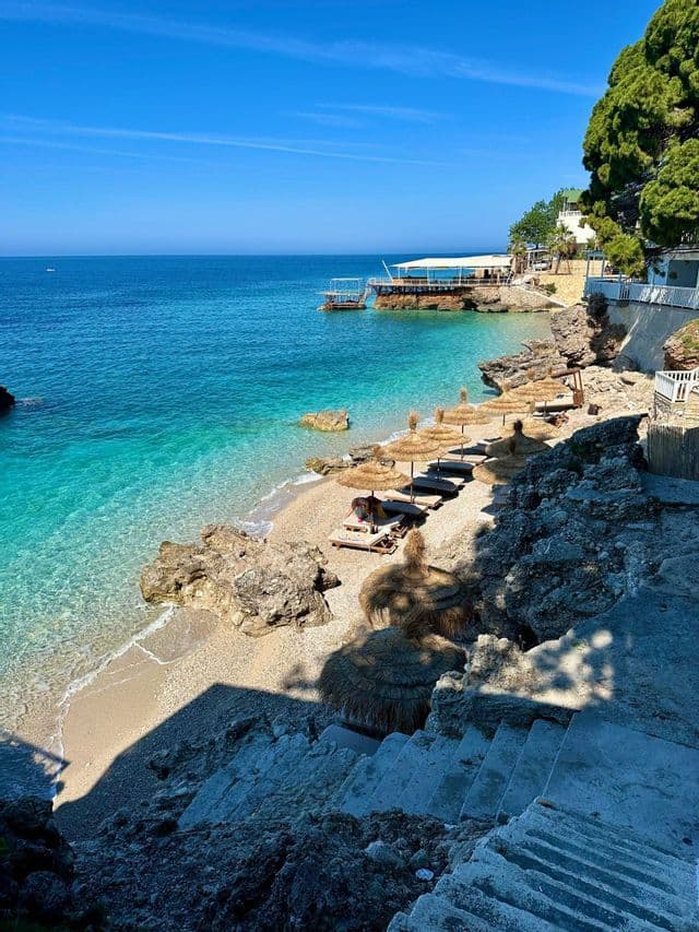 Una vista elevada desde escaleras de piedra que dan a una playa rocosa y apartada con sombrillas de paja y tumbonas junto a aguas turquesas.
