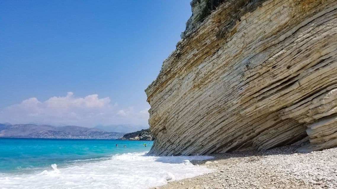 Un gran acantilado de roca estratificada se une a una playa de guijarros donde el agua turquesa con suaves olas lame la orilla bajo un cielo azul claro.