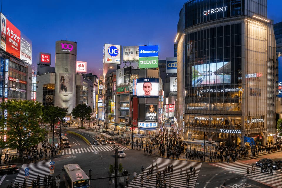 Una vasta folla di persone attraversa un trafficato incrocio cittadino al tramonto, circondata da alti edifici con luminose insegne al neon.