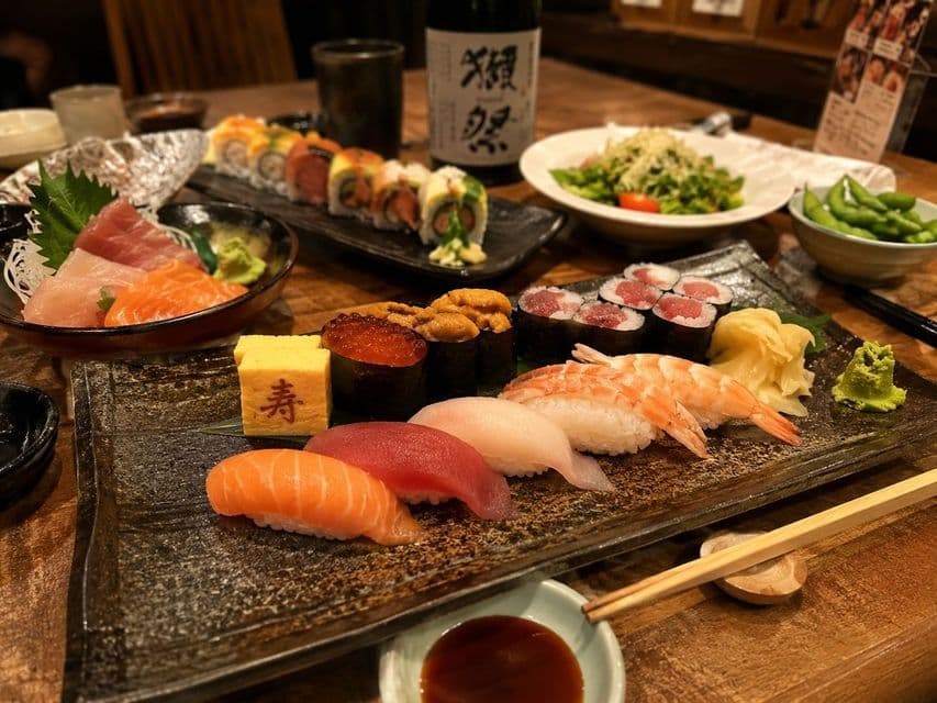Piatti assortiti di sushi, sashimi e roll sono disposti su un tavolo di legno con contorni e una bottiglia di sake.