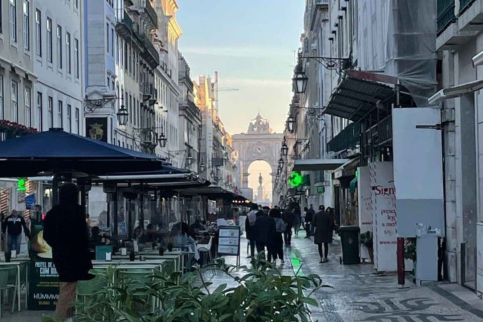 Una strada cittadina fiancheggiata da edifici e caffè all'aperto, con persone che camminano verso un grande arco alla fine.