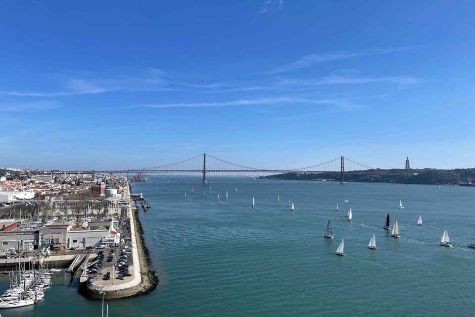 Veduta aerea di barche a vela su un ampio fiume, con un grande ponte sospeso rosso e una città costiera sullo sfondo.