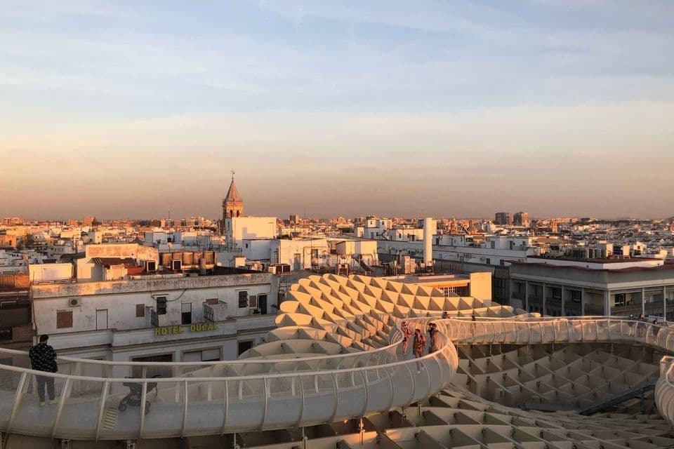People walk on a curving walkway over a modern honeycomb structure, with a cityscape and church tower visible at sunset.
