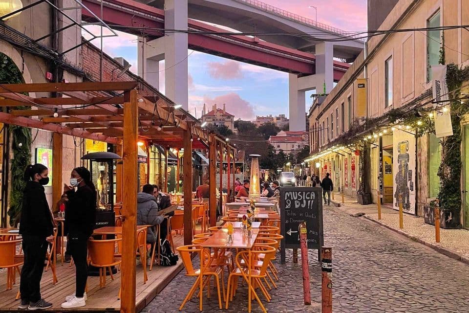 Un ristorante all'aperto su una strada di ciottoli con persone che cenano sotto un grande ponte al tramonto.