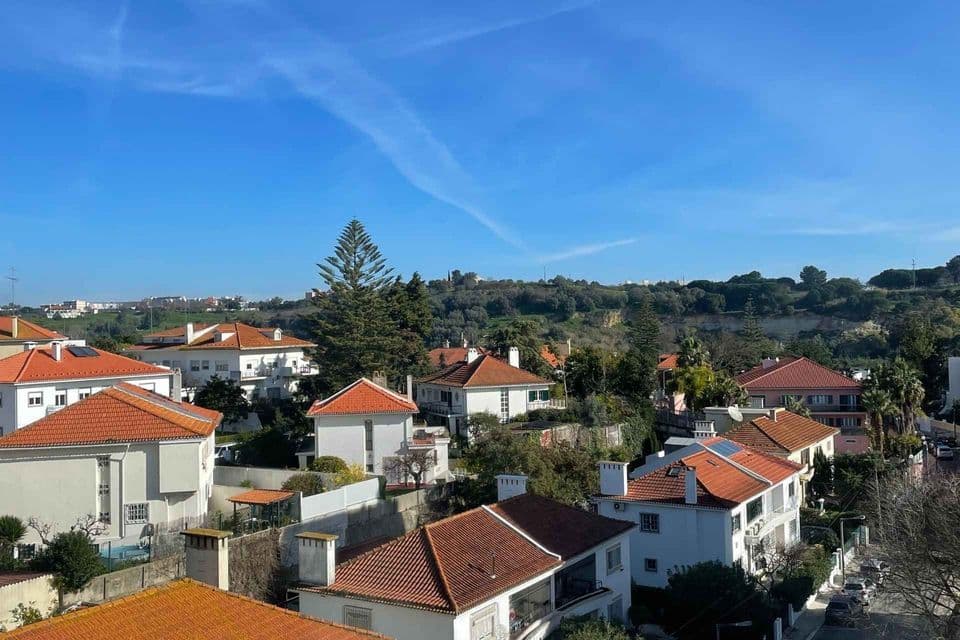 Una vista dall'alto di case dai tetti rossi sparse su un fianco di collina verde e alberato sotto un limpido cielo azzurro.