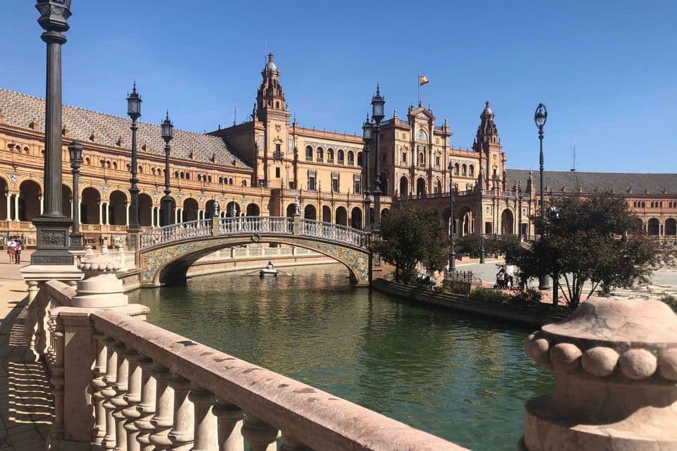 A grand, arched building with a decorative bridge over a canal, where a person is rowing a small boat under a clear blue sky.