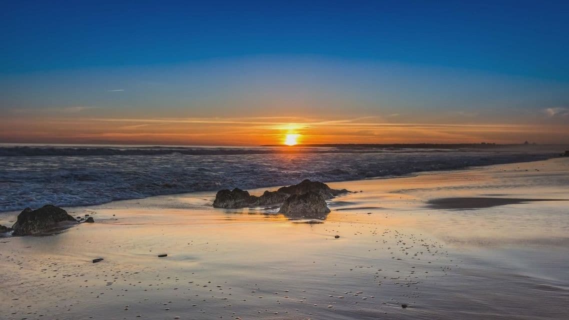 Il sole tramonta sull'oceano, con le onde che lambiscono una spiaggia di sabbia bagnata disseminata di rocce scure sotto un cielo blu e arancione.