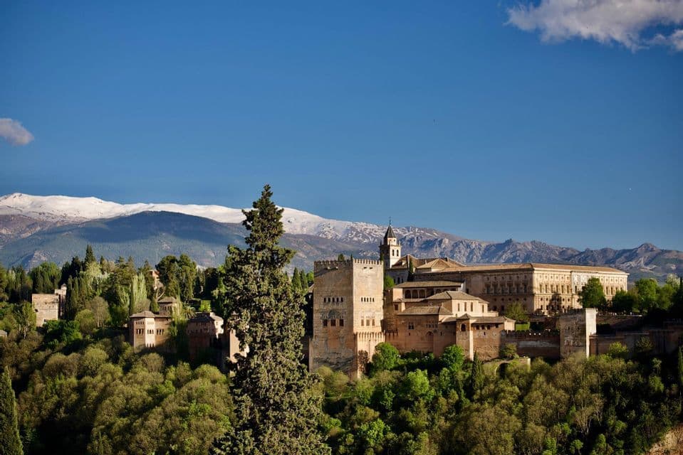 Una storica fortezza di pietra su una collina boscosa con uno sfondo di montagne innevate sotto un cielo azzurro e limpido.