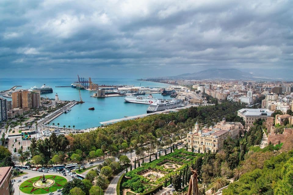 Una veduta aerea di una grande città costiera con un porto trafficato, navi da crociera e rigogliosi parchi verdi sotto un cielo nuvoloso.