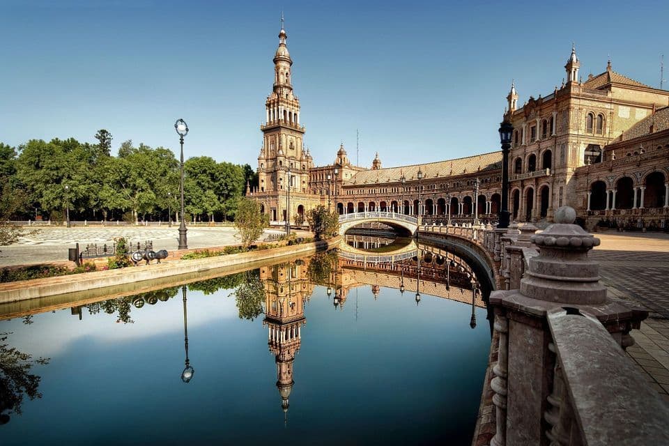 Un edificio sontuoso con un'alta torre si riflette nell'acqua calma di un canale sinuoso sotto un cielo azzurro e sereno.