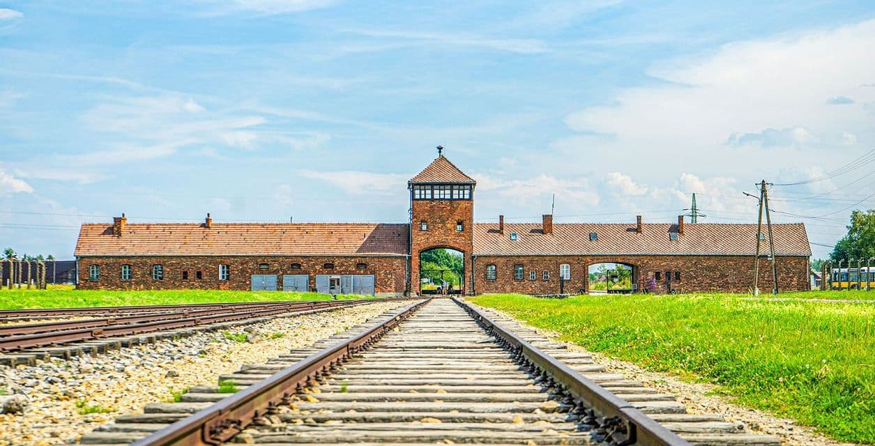 Binari ferroviari conducono a un'imponente portineria in mattoni con una torre di guardia centrale, sotto un cielo soleggiato e nuvole sparse.
