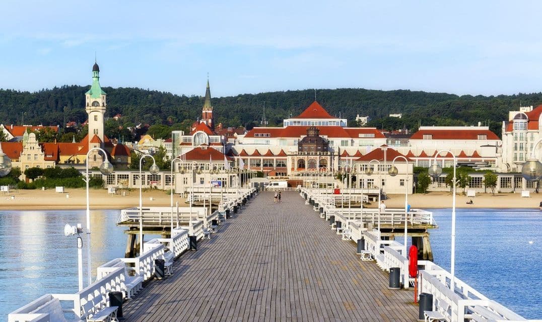Un lungo pontile in legno con ringhiere bianche conduce verso una cittadina costiera in stile europeo con una spiaggia sabbiosa e una collina boscosa sullo sfondo.