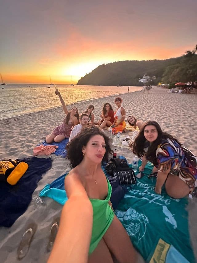 Une femme prend un selfie de son groupe WeRoad lors d'un pique-nique sur une plage de sable, au coucher du soleil coloré.