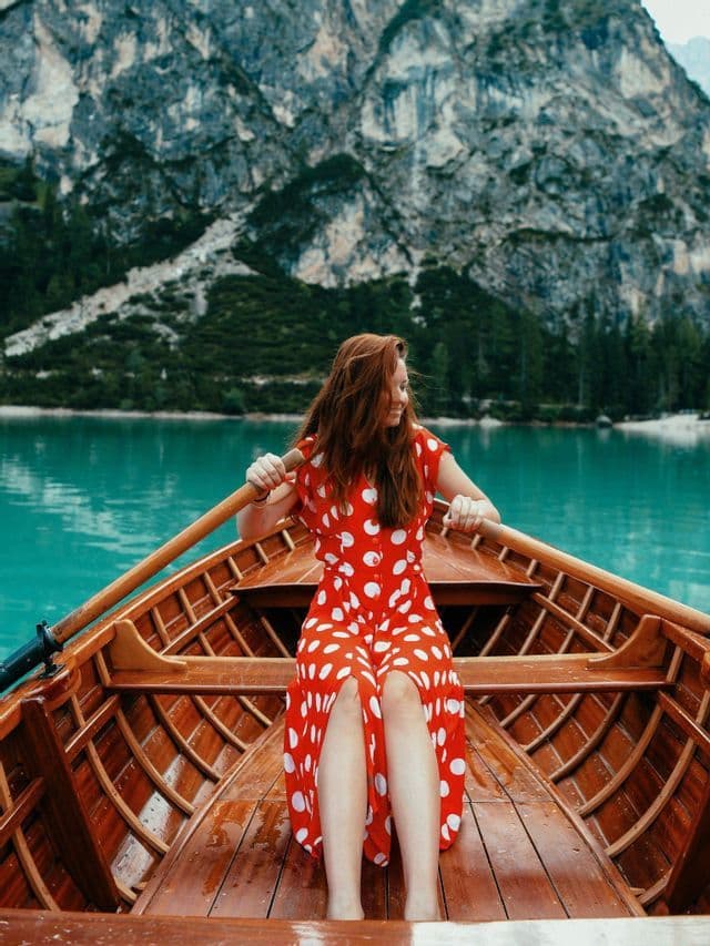 Eine Frau in einem roten Tupfenkleid rudert ein Holzboot auf einem türkisfarbenen See mit Bergen im Hintergrund.
