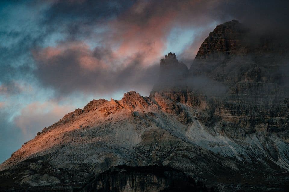 Zerklüftete Berggipfel im warmen Sonnenuntergangslicht unter dunklen, farbenfrohen Wolken und Nebel.